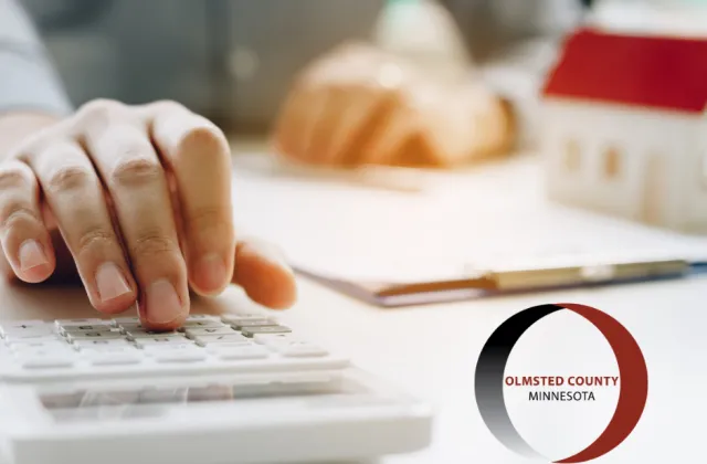 An individual is using a calculator with documents and a small model house on a desk, next to a logo of Olmsted County, Minnesota.