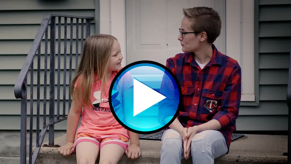 Adrian and their daughter sitting on a step. Play button in the middle of the photo