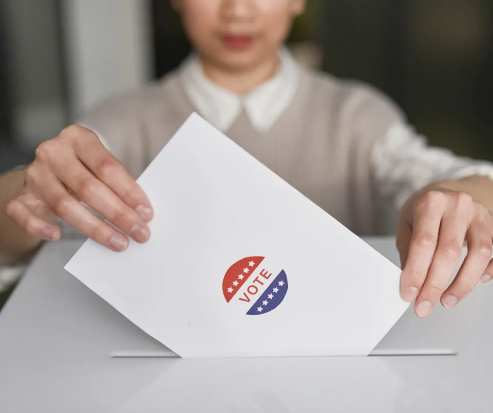 A person putting a ballot in a box. The ballot has a sticker on it that says, "VOTE."