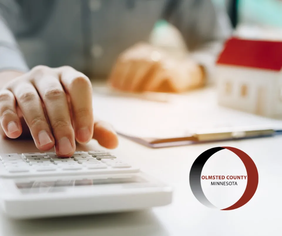 An individual is using a calculator with documents and a small model house on a desk, next to a logo of Olmsted County, Minnesota.