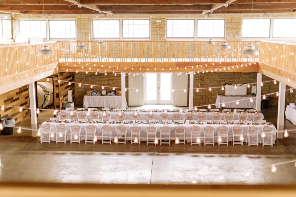 Interior of a rustic event hall with exposed brick walls and wooden beams, decorated with string lights. Tables are set with white linens and chairs arranged for a large gathering.