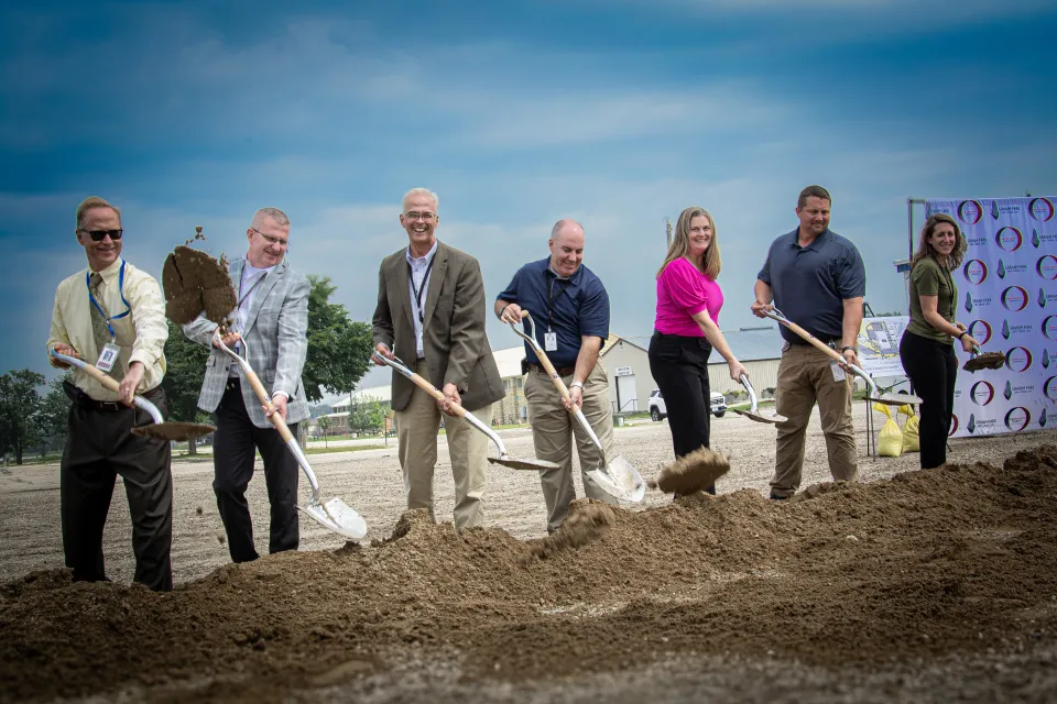 A group of seven people, dressed in business and casual attire, participate in a ceremonial groundbreaking event. Each person is smiling and holding a shovel, tossing dirt into the air. They stand in a row on a dirt lot under a partly cloudy sky, with buildings and event signage visible in the background.