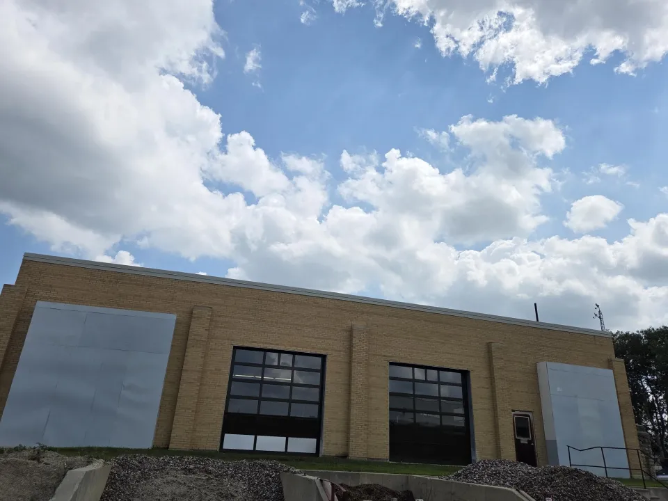 A low-angle view of a tan brick building with two large black-framed garage doors and metal panels covering parts of the facade. A single brown door with a small awning is visible on the right. The sky above is bright and partly cloudy, and construction materials like gravel and dirt are seen in the foreground, indicating ongoing work.