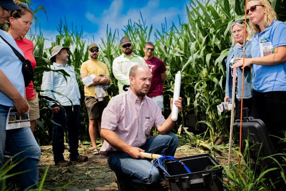 Man holding an instrument to measure the quality of soil. A cornfield and a group of people stand in the background.