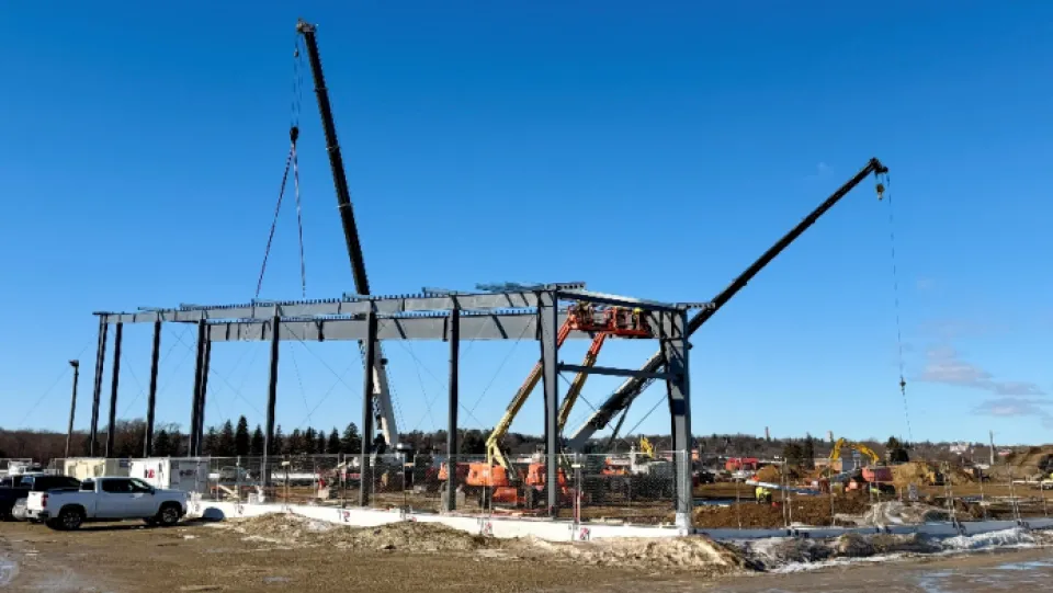 Construction site with steel framework partially erected. Multiple cranes are in operation, lifting beams. Machinery and construction vehicles are visible around the site. Clear blue sky and some snow on the ground.