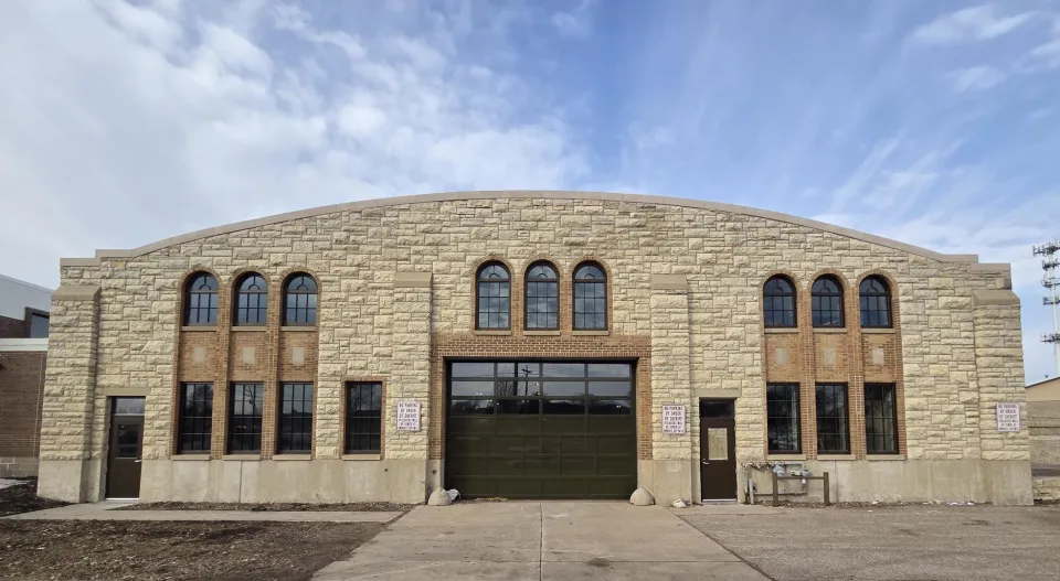 A large stone building with multiple arched windows and a central garage door. The facade features various stone textures, and a clear sky with light clouds is in the background.