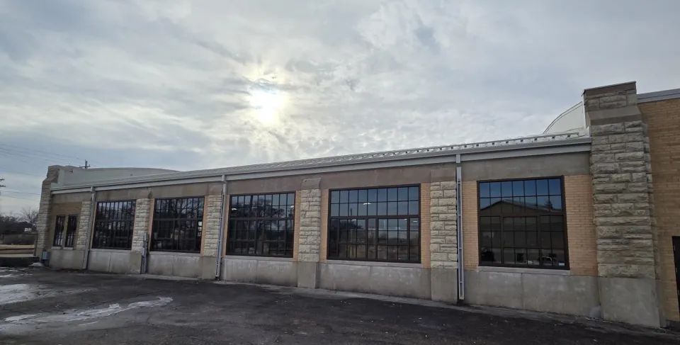 A large industrial-style building with multiple tall windows and stone accents under a cloudy sky. The ground appears to be a paved outdoor space, and the sun is partially visible, casting a soft light over the scene.