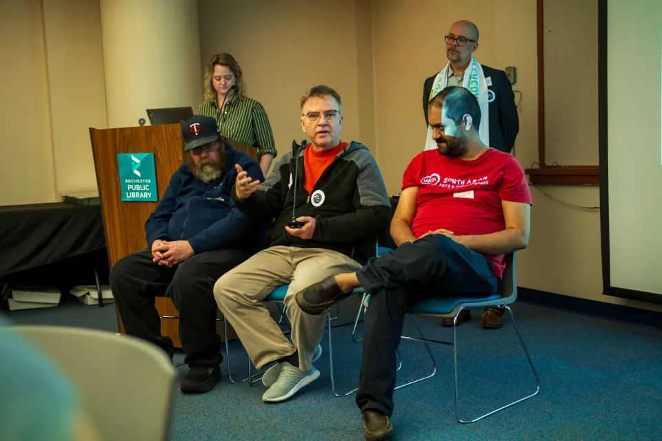 Three men sit at the front of a room in a row. The man in the middle is holding a microphone and talking