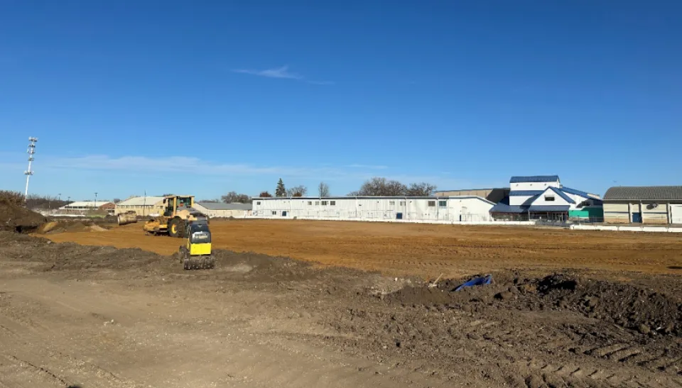 A construction site with earthmoving equipment working on a large dirt field. In the background, there are several white industrial buildings and one building with blue accents. The sky is clear and blue.