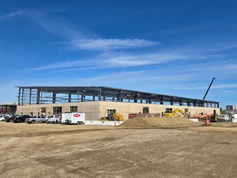 A large construction site with a partially built structure, featuring steel frames and a beige facade. Several construction vehicles and workers are present around the building. A crane is positioned on the right. The sky is clear and blue.