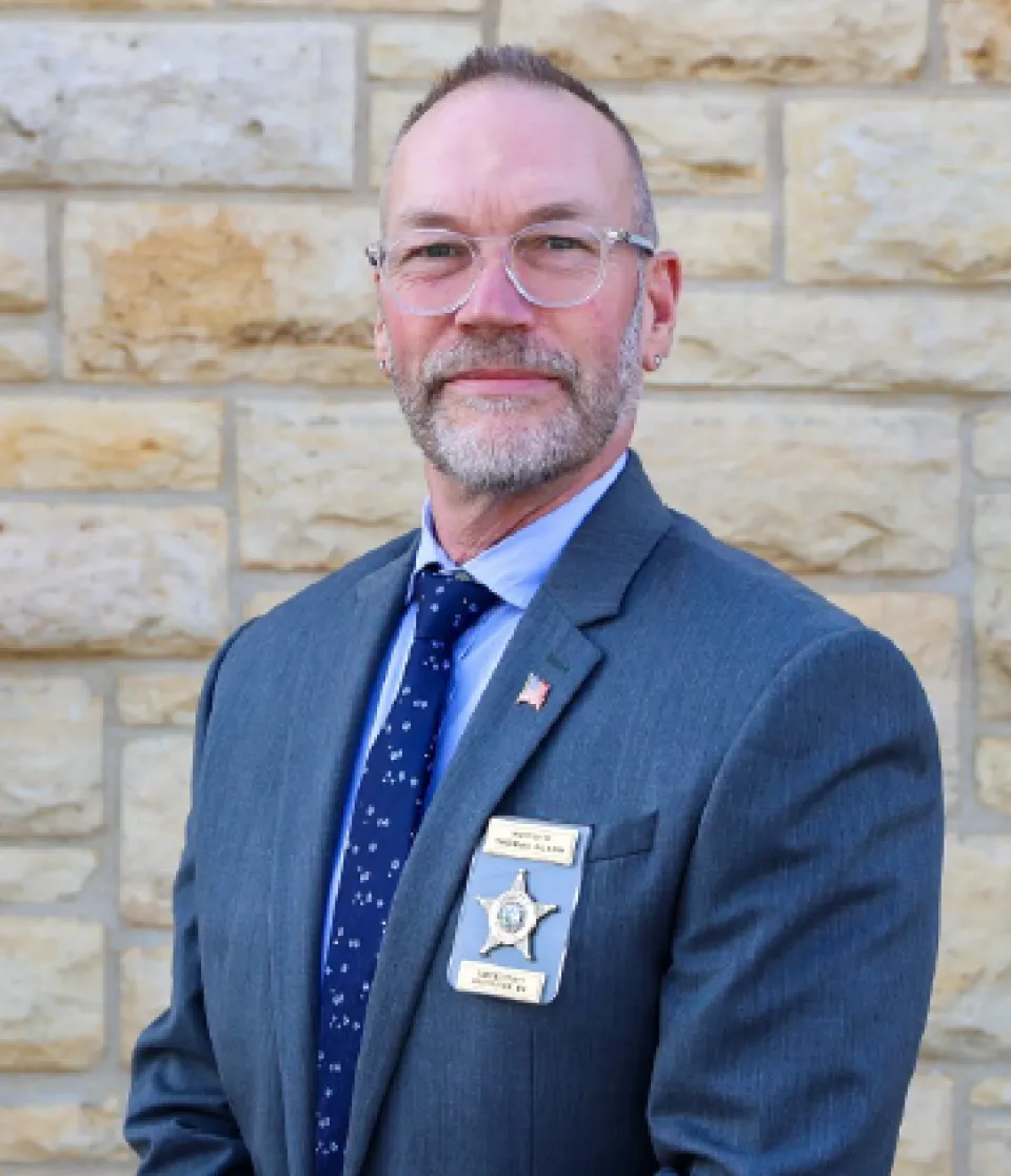 Tom Olson head shot wearing a suit and standing in front of a stone building. 