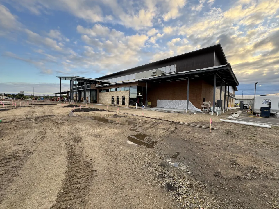 A large industrial building under construction with a modern design is shown. The structure features a mix of brick and metal siding. The foreground includes dirt and construction materials, with scattered puddles from recent rain. The sky above is partly cloudy.