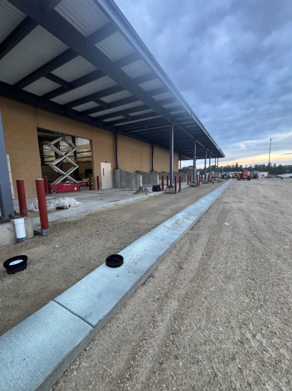 Construction site with a large building under development. The building features a metal roof supported by beams and has an unfinished facade. Scattered construction materials and equipment are visible. The ground is covered with gravel, and the sky is overcast.