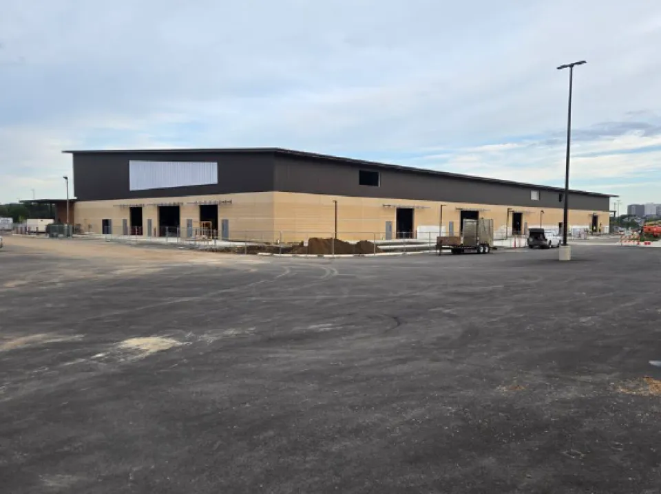 A large, newly constructed industrial building with a modern design, featuring a beige and dark grey exterior. The surrounding area is paved and mostly empty, with some construction materials and equipment visible near the building. The sky is partly cloudy.