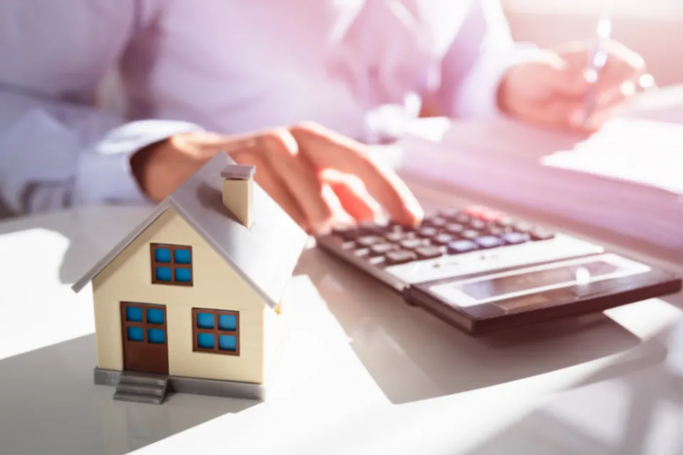 A person touching a calculator with a small, model house in the forefront.