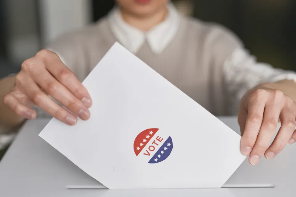 A person putting a ballot in a box. The ballot has a sticker on it that says, "VOTE."