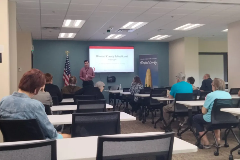 Olmsted County PRL Associate Director Luke Turn gives a presentation about the ballot board process in Olmsted County. Several people sitting at tables, watching and listening to the presentation. 