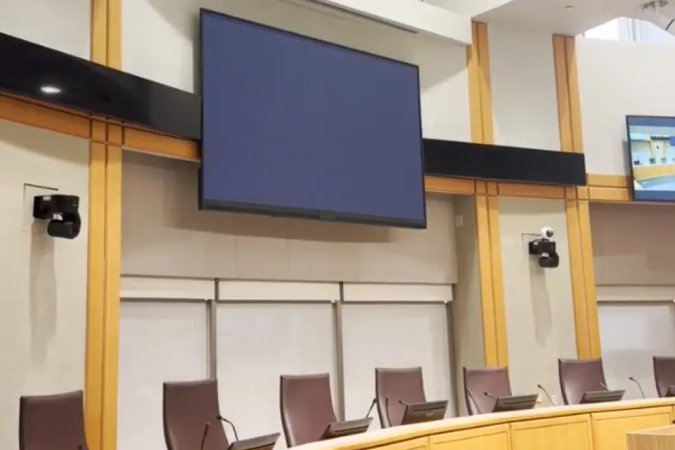 A modern conference room featuring a large blank screen at the front, surrounded by wood-paneled walls and several rows of empty chairs facing the screen.