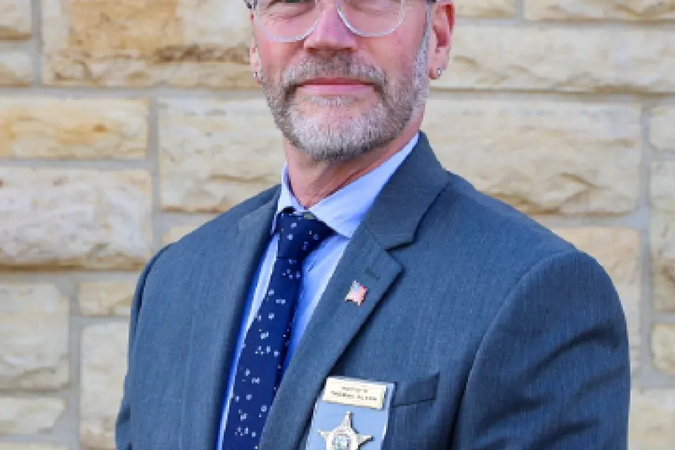 Tom Olson head shot wearing a suit and standing in front of a stone building. 